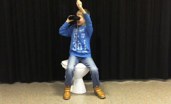 Ein junge sitzt vor einem Vorhang auf einer Toilette mit runtergeklapptem Deckel, schaut durch ein Fernglas und hat die Hand in Siegerpose erhoben.