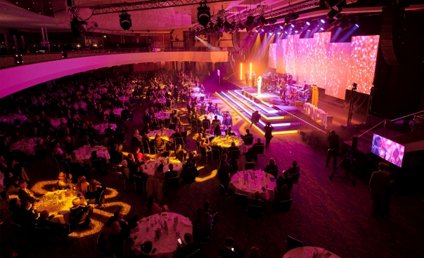Seitlicher Blick auf einen Festsaal voller runder Tische und mit Bühne, auf der Programm stattfindet.