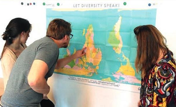 Drei Personen schauen sich eine Weltkarte an. Ein Person zeigt auf ein afrikanisches Land.
