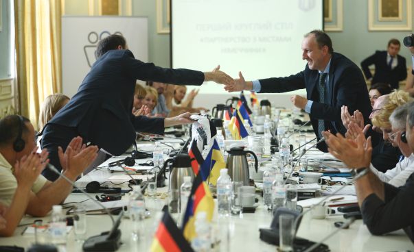 Personen sitzen an einem Konferenztisch, auf dem ukrainische und deutsche Flaggen stehen.