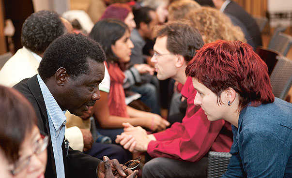 An African Man and a white woman with short red hair are sitting in front of each other and talking between lots of other people.
