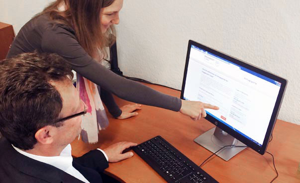 Ein Mann sitzt vor dem Computer und eine Frau steht neben ihm und zeigt auf den Bildschirm, auf dem man das Antragsportal sieht.