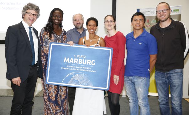 Die Gewinner Marburg des Wettbewerbs „Kommune bewegt Welt“ stehen für das Gewinnerfoto zusammen und halten ein blaues Ortsschild hoch. Foto: Tobias Vollmer