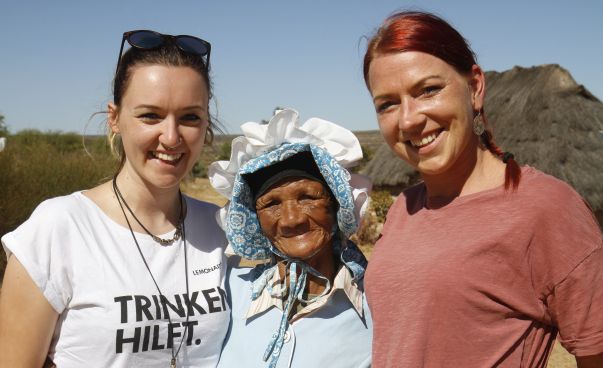 Eine ältere Frau steht mittig zwischen zwei jungen Frauen. Sie lächeln in die Kamera.
