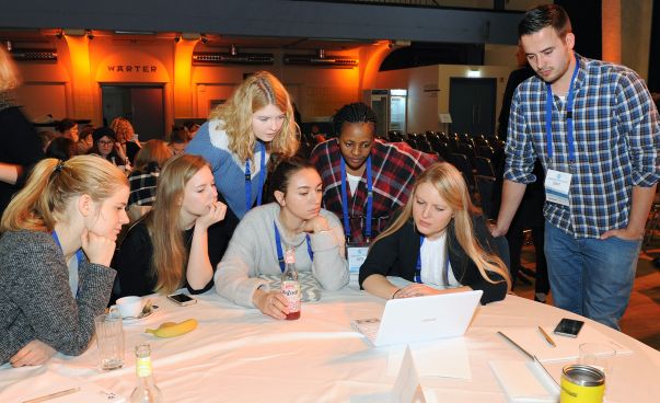 Eine Gruppe von Menschen verschiedener Herkunft sitzt zusammen an einem runden Tisch und schaut gemeinsam auf einen Laptop. Foto: Bodo Tiedemann/Engagement Global
