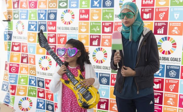 Eine junge Frau und ein Mädchen stehen vor einer mit den Nachhaltigkeitszielen bedruckten Wand. In den Händen halten Sie bunte Gegenstände. Foto: Engagement Global