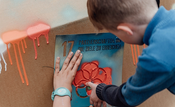 Ein Junge sprüht Farbe auf eine Schablone die eines der Nachhaltigkeitsziele darstellt. Foto: Engagement Global/Charlotte Wulff