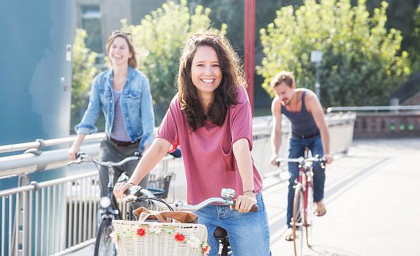 Eine Frau fährt auf einem Fahrrad und blickt lächelnd in die Kamera. Hinter ihr sind zwei weitere Personen auf Fahrrädern. Die Sonne scheint.
