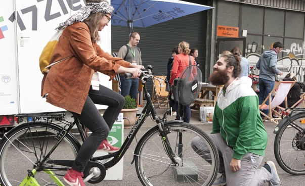 Eine Frau fährt auf einem fixierten Fahrrad; es ist an einen Ventilator angeschlossen. Ein Mann kniet vor dem Ventilator. Foto: Jo Hempel