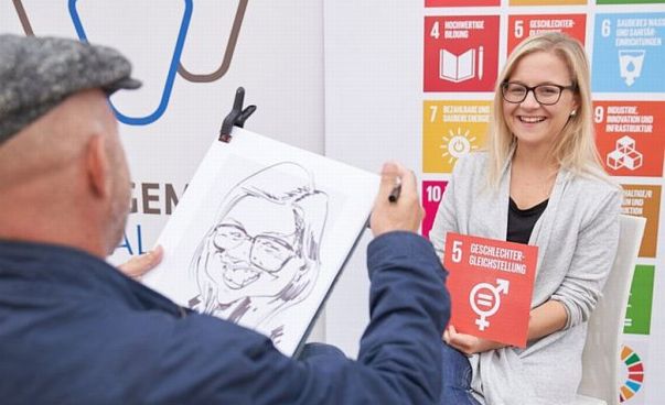 Eine Frau hält ein Schild mit einem Nachhaltigkeisziel in der Hand. Es zeigt Ziel 5 Geschlechtergereichtigkeit an. Sie wird von einem Karikaturisten gezeichnet.