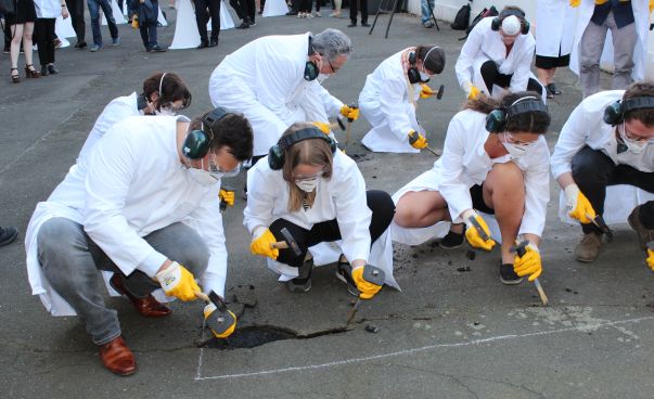 Eine Gruppe von Menschen in weißen Kitteln entsiegeln eine asphaltierte Fläche mit Hammer und Meißel. Foto: Engagement Global