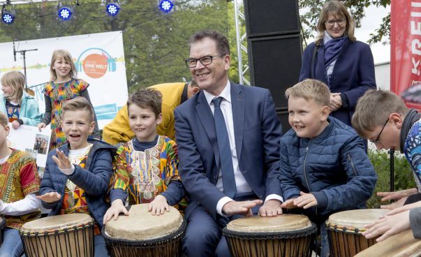 Bundesminister Gerd Müller sitzt neben Schülern beim Tag der offenen Tür. Die Kinder trommeln auf Bongos. Foto: Engagement Global/Merlin Nadj-Torma