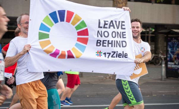 Zwei Männer halten während sie laufen ein Plakat in die Höhe mit dem SDG Rad sowie dem Spruch "Leave no one behind #17Ziele"
