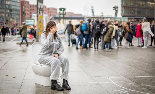 Eine Schülerin sitzt in einem Overall auf einer Kloschüssel mitten auf dem Potsdamer Platz in Berlin. Foto: Delia Wöhlert