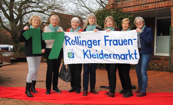Sieben Organisatorinnen des Frauen-Kleidermarktes halten ein Schild mit der Aufschrift „17. Rellinger Frauen-Kleidermarkt“ in die Kamera. Foto: Stefan Flomm