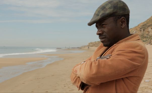 Ein Mann steht mit Schiebermütze und Lederjacke und verschränkten Armen am Strand und schaut in Richtung Meer. Foto: jip Film