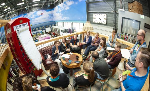 Eine Gruppe von rund 15 Menschen unterschiedlichen Alters sitzt um einen kleinen runden Holztisch herum auf einer Empore im Basecamp Bonn. Die Menschen diskutieren und halten Ideen  schriftlich auf einem Flipchart fest. Foto: Kolja Matzke