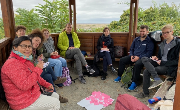 Eine Gruppe von Menschen sitzt in einer runden Ausflugshütte. In ihrer Mitte liegt ein weißes Poster mit pinken Post-its.
