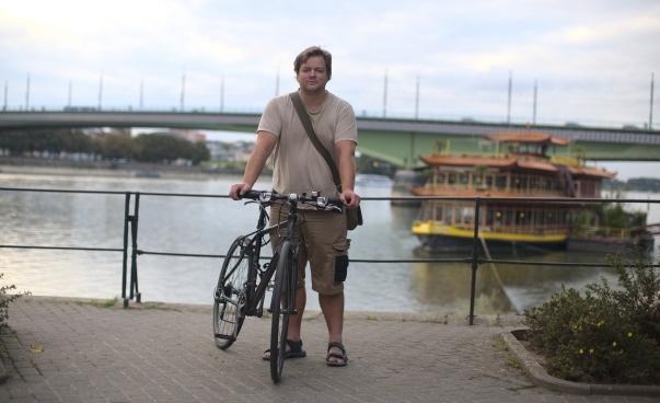 Ein Mann mit Fahrrad steht vor dem Rhein, im Hintergrund ist ein Ausflugsschiff zu sehen.