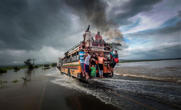 Ein Bus mit vielen Menschen fährt über eine überschwemmte Straße.
