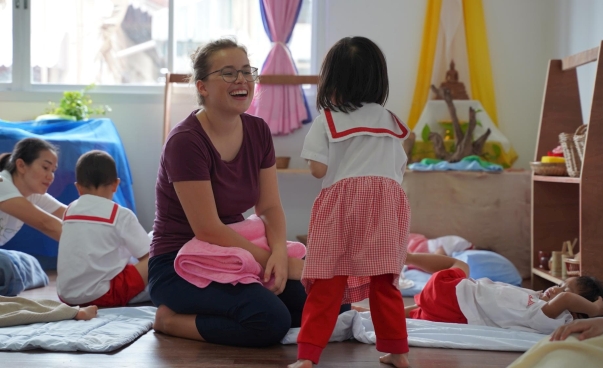 Eine junge Frau lacht in einer Kindertagesstätte mit einem kleinen Mädchen. Im Hintergrund spielen weitere Kinder.