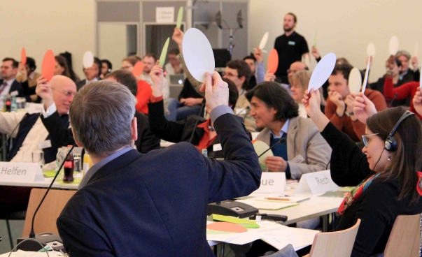 Eine Gruppe von Menschen in einem Konferenzraum stimmt mit erhobenen bunten Zetteln ab.