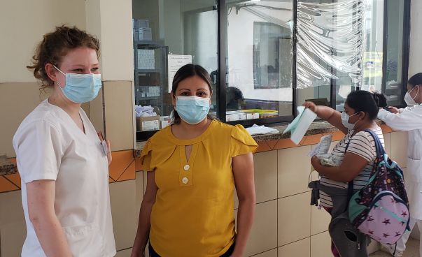 Zwei Frauen, eine davon in medizinischer Kleidung, posieren für ein Foto. Im Hintergrund die Anmeldestation eines Krankenhauses, wo zwei andere Frauen Formulare ausfüllen.