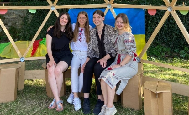 Vier Mädchen sitzen nebeneinander auf einer Bank in einer Holzkonstruktion. Im Hintergrund eine ukrainische Flagge.