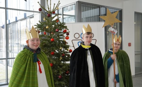 Drei Kinder in Königskostümen vor einem Weihnachtsbaum mit Spendenbox in der Hand.