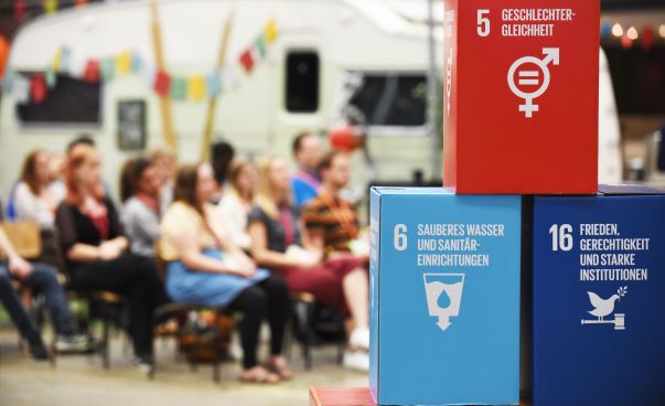 Würfel mit drei der 17 SDG Symbolen. Im Hintergrund eine Gruppe sitzender Menschen und ein Wohnwagen.