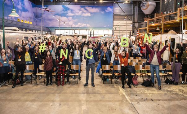 Eine große Gruppe von menschen steht in einer Halle und lächelt in die Kamera. Die Personen in der ersten Reihe halten die grünen Buchstaben #NCBN in die Luft.