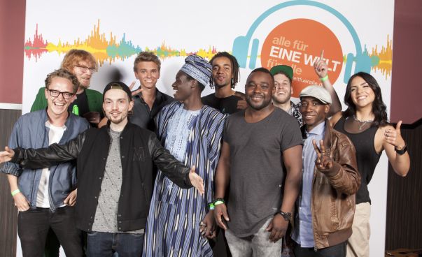 Eine Gruppe junger Menschen steht vor dem Plakat des Eine Welt Song Contest.