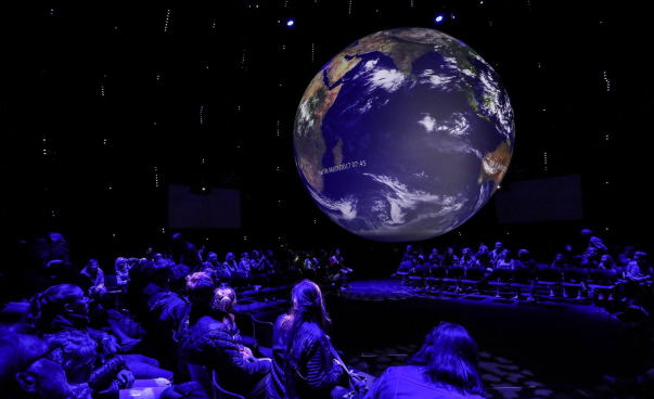 Eine beleuchtete Weltkugel hängt in einem dunklen Raum. Um sie herum sitzt Publikum und betrachtet sie. Foto: Global Citizen