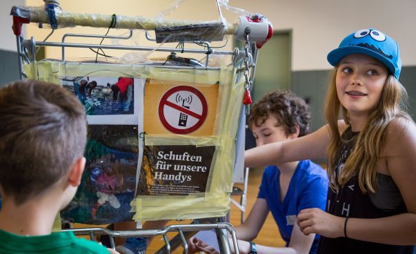 Ein Mädchen sitzt vor einem Einkaufswagen, der mit bunten Zeitungsausschnitten beklebt ist. Darauf ist ein durchgestrichenes Handy und darunter die Schrift "Schuften für unsere Handys" abgebildet. Foto: Anja Lehmann