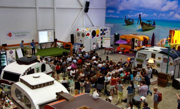 In einer großen bunten Halle mit Wohnwagen stehen die Teilnehmenden des NachhaltigkeitsCamps 2016 zusammen.