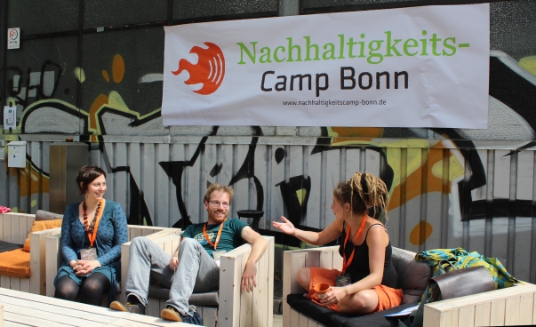 Drei Menschen sitzen draußen vor einer Halle und diskutieren. Im Hintergrund hängt ein Banner vom NachhaltigkeitsCamp Bonn.