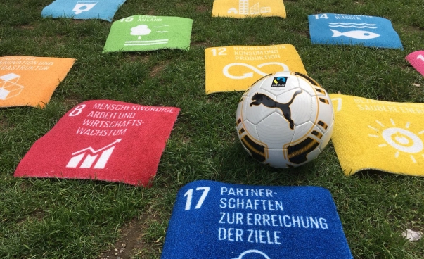 Mehrere Teppiche mit Aufdruck der Ziele für nachhaltige Entwicklung liegen um einen Fußball auf Rasen.