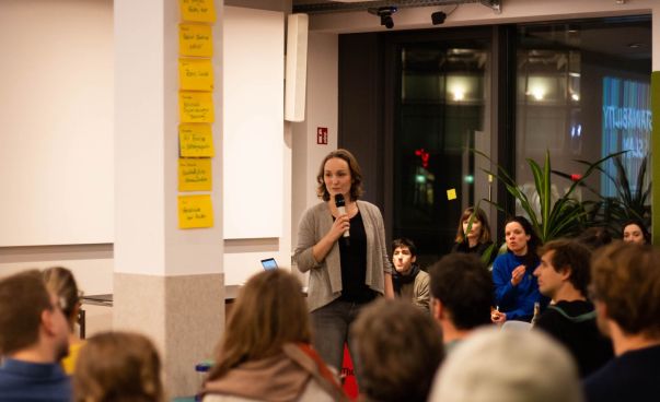 Eine Frau steht mit einem Mikrofon vor einem Publikum und spricht. Foto: Nico Scagliarini
