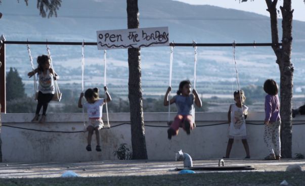 Vier Kinder schaukeln. Im Hintergrund ist das Meer zu sehen. Auf einem Schild steht open the borders.