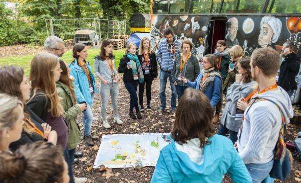 Eine Gruppe junger Menschen hat sich unter freien Himmel im Kreis um eine Weltkarte versammelt. Im Hintergrund steht ein bemalter Bus.