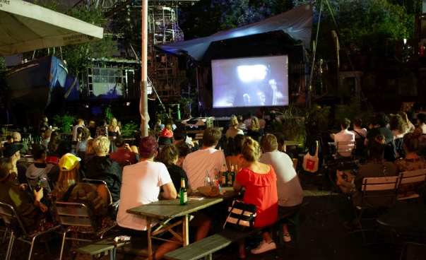 Eine große Gruppe von Menschen sitzt im Freien und schaut auf eine Leinwand, auf der ein Film gezeigt wird.