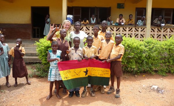 Eine Gruppe Kinder steht mit einer Freiwilligen von weltwärts vor einer Schue. Sie halten die Flagge Ghanas vor sich.