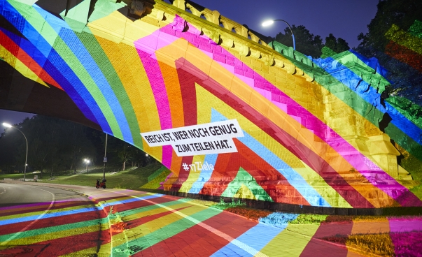 Brücke mit Textprojektion in SDG-Farben: Reich ist, wer noch genug zum Teilen hat.