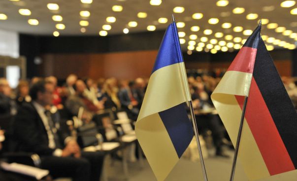 Die ukrainische und deutsche Flagge stehen auf einem Tisch. Im Hintergrund sind Menschen zu sehen.