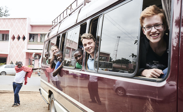 Fünf Jugendliche schauen lachend hintereinander aus einem roten Kleinbus aus den Fenstern. Neben dem Bus steht ein Jugendlicher mit der Hand an den Bus gelehnt.