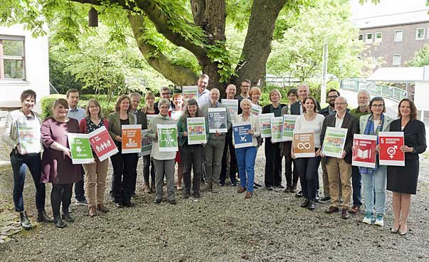 enschen halten Schilder mit den Symbolen der 17 nachhaltigen Entwicklungsziele im Halbkreis, im Hintergrund steht ein großer Baum.