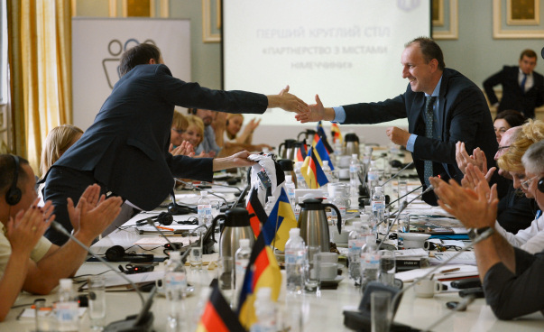 Zwei Männer geben sich die Hand und beugen sich über einen Tisch. Auf dem Tich sind deutsche und ukrainische Fahnen zu sehen, im Hintergrund ist eine Präsentation an die Wand geworfen. Foto: Jaroslaw Kostyk